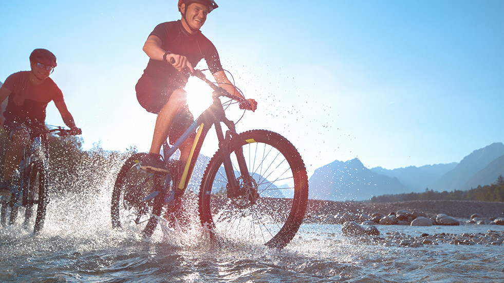 Zwei Mountainbiker fahren durch flaches Wasser, spritzendes Wasser, Berglandschaft im Hintergrund