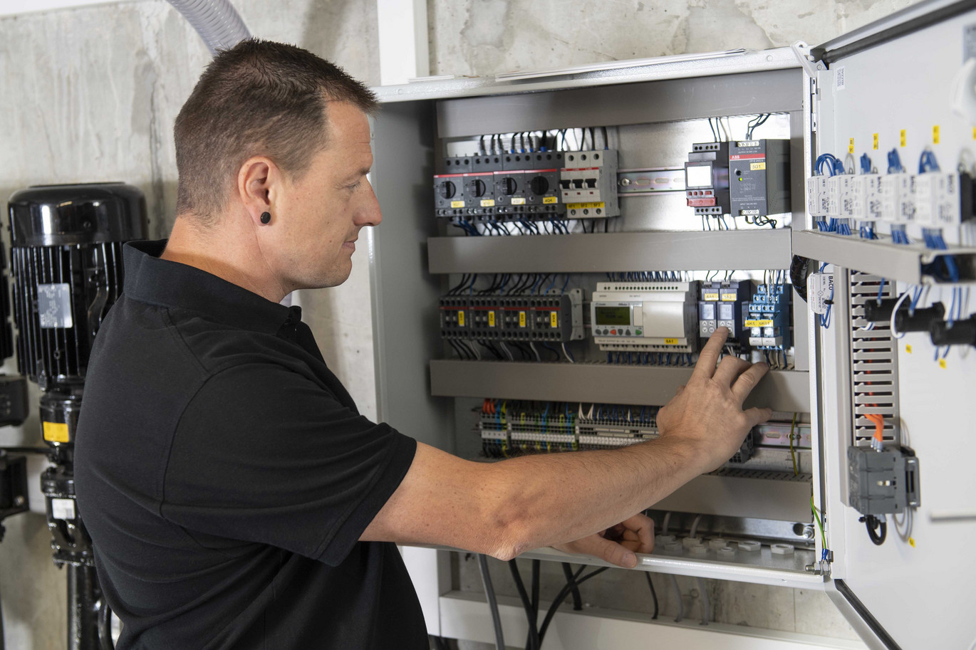 Ein Techniker arbeitet konzentriert an den elektrischen Komponenten eines Schaltschrankes, umgeben von Kabeln und technischen Geräten im Hintergrund