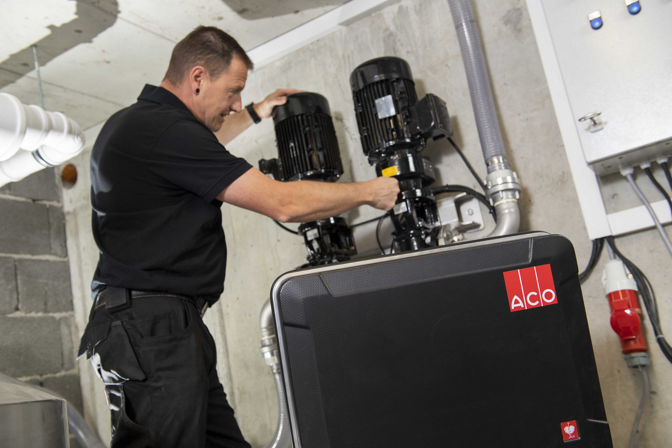 Ein Techniker arbeitet an zwei großen schwarzen Pumpen in einem technischen Raum. Im Vordergrund ist ein schwarzes Gerät mit dem ACO-Logo zu sehen