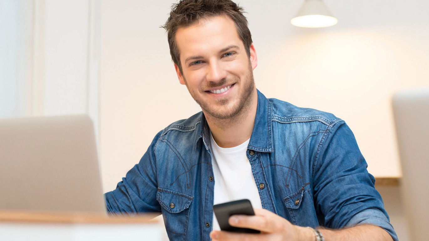 Mann in Jeanshemd lächelt und hält ein Smartphone, sitzt vor einem Laptop in einem hellen Büro