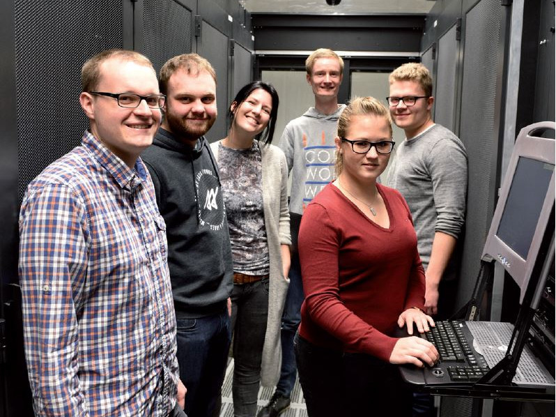 Sechs lächelnde Menschen stehen in einem Serverraum, eine Frau arbeitet an einem Computer