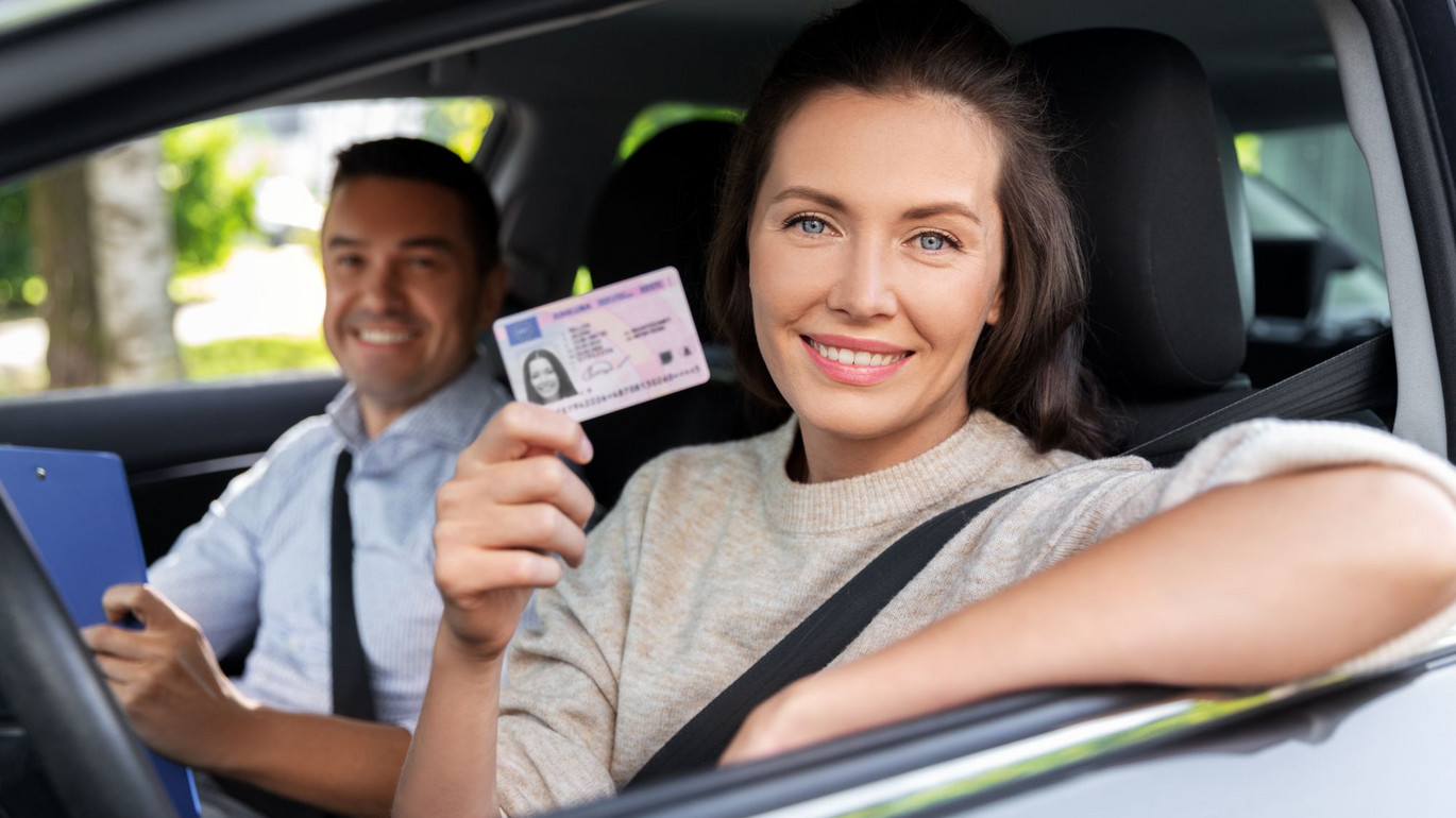 Frau im Auto hält ihren Führerschein, lächelt, Fahrlehrer sitzt daneben