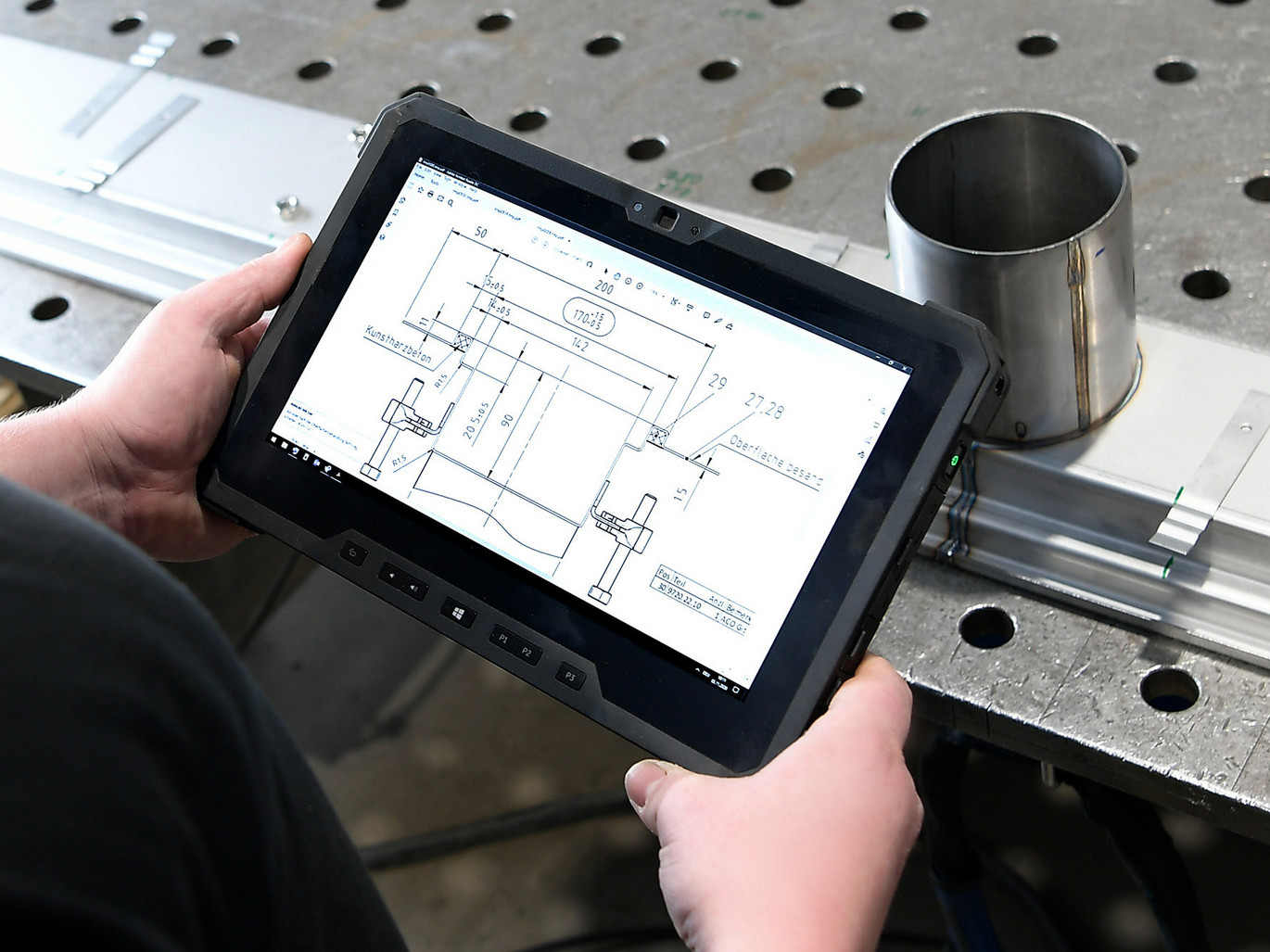 Person hält ein Tablet mit technischer Zeichnung in einer industriellen Umgebung, Metallarbeitstisch und Metallrohr im Hintergrund.