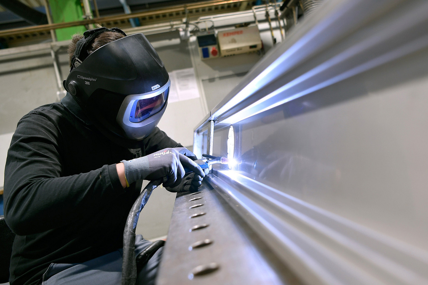 Person mit Schweißhelm und Schutzhandschuhen schweißt ein Metallstück in einer Werkstatt, während helles Licht den Arbeitsbereich beleuchtet
