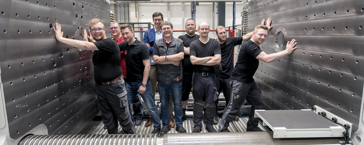 Eine Gruppe von neun Männern in Arbeitskleidung posiert lächelnd zwischen großen industriellen Maschinen in einer Werkshalle